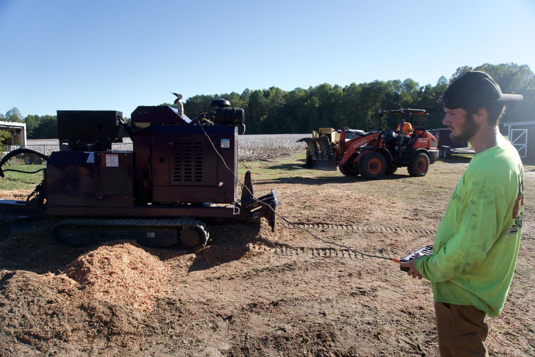 Tree Works crew member running a remote-controlled tracked stump grinder at the edge of a cotton field