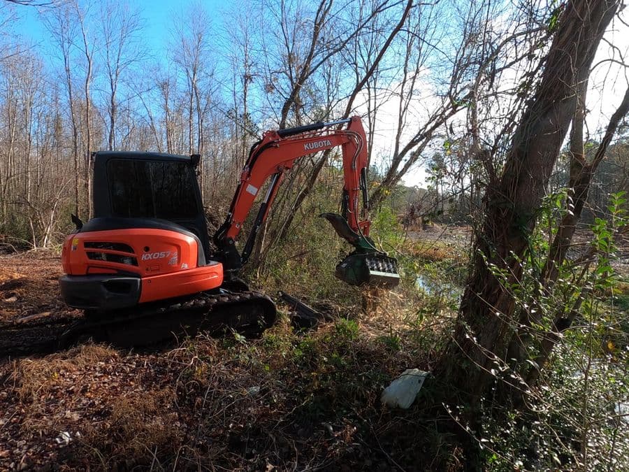 Kubota excavator with a forestry mulching head clearing brush along a creek